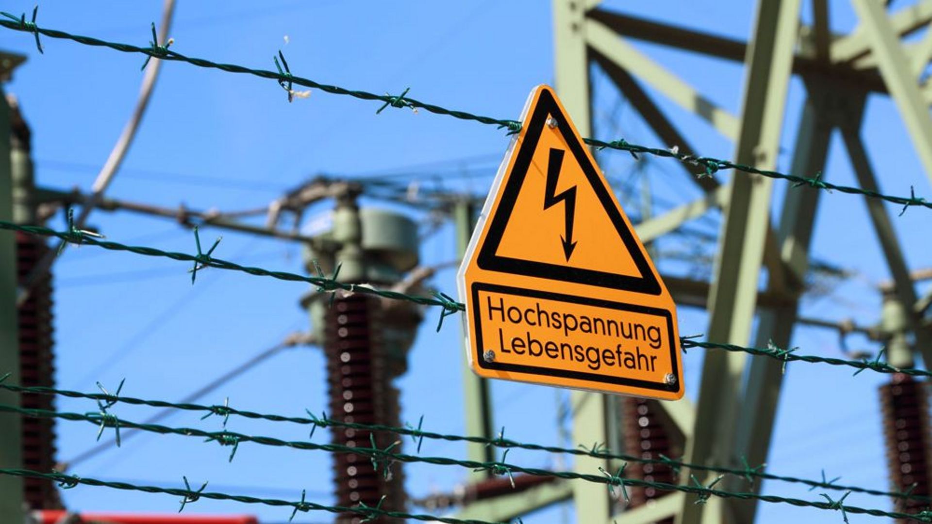 In einer Trafostation ist ein Schild befestigt. Ein orangenes Dreieck mit schwarzer Kontur und einem schwarzen Blitz in der Mitte. Darunter befindet sich ein Rechteck mit der Aufschrift "Hochspannung Lebensgefahr"
