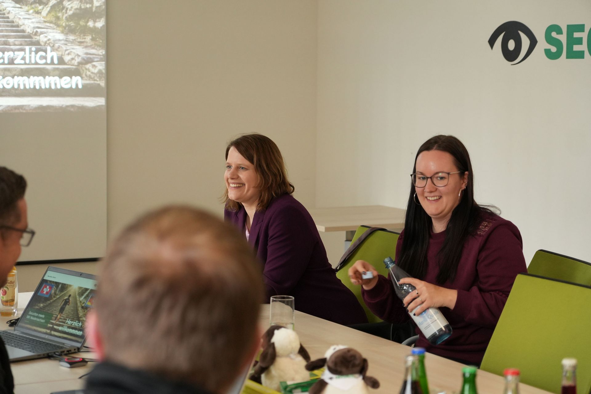Die stellvertretende niedersächsische Ministerpräsidentin Julia Willie Hamburg sitzt in einem Besprechungsraum an einem Tisch und lächelt. Neben ihr sitzt eine Mitarbeiterin, die eine Wasserflasche hält. Auf dem Tisch stehen Getränke und mehrere kleine Stofftiere. Im Hintergrund ist eine Präsentationsfolie teilweise sichtbar.