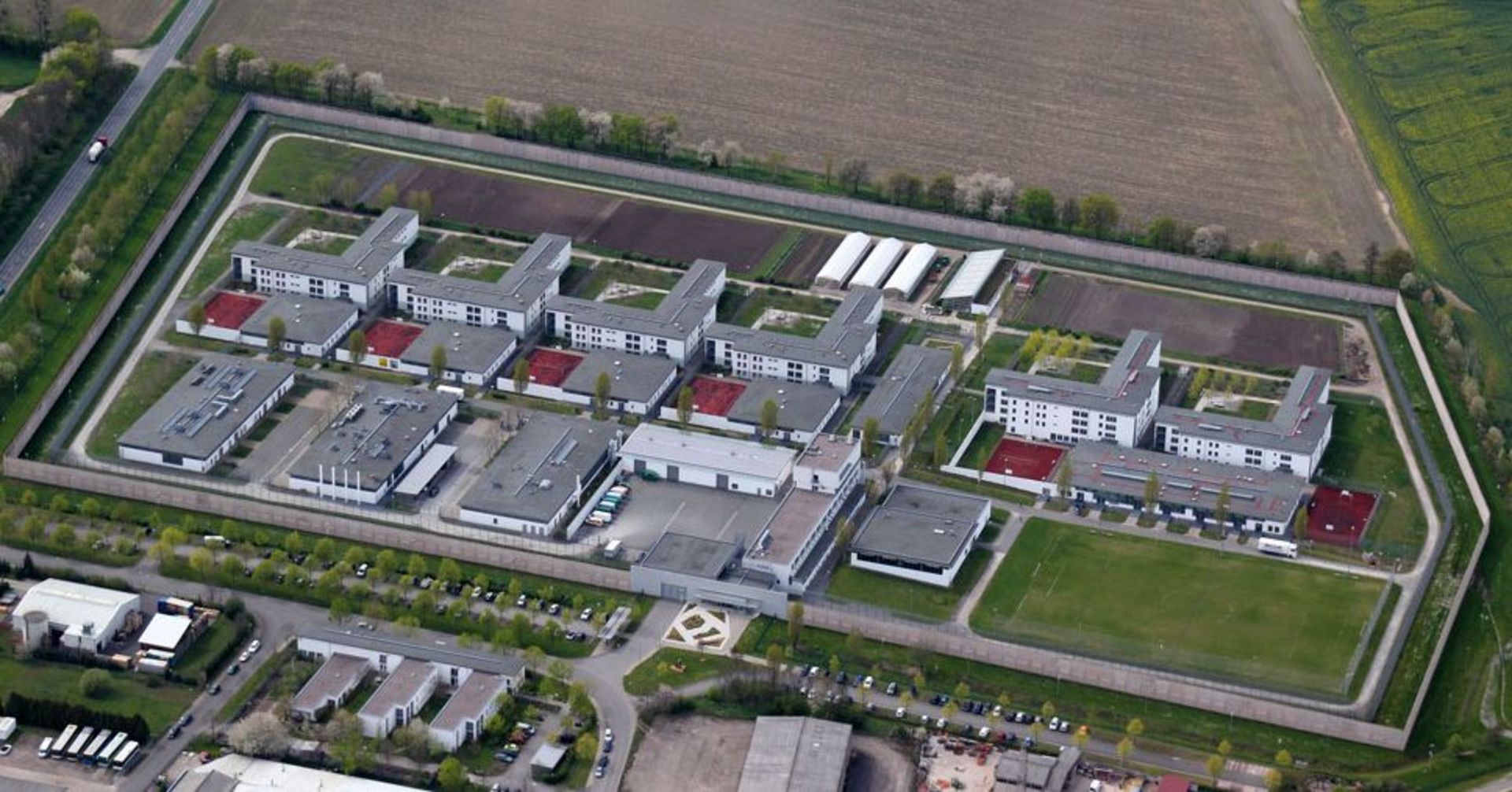 Luftaufnahme eines weitläufigen, rechteckig ummauerten Justizvollzugsanstalten Geländes mit mehreren Gebäudekomplexen, Innenhöfen, Sportflächen und Wirtschaftsbereichen. Das Areal ist vollständig von einer hohen Sicherheitsmauer umgeben und von landwirtschaftlichen Flächen sowie Gewerbegebäuden eingerahmt.