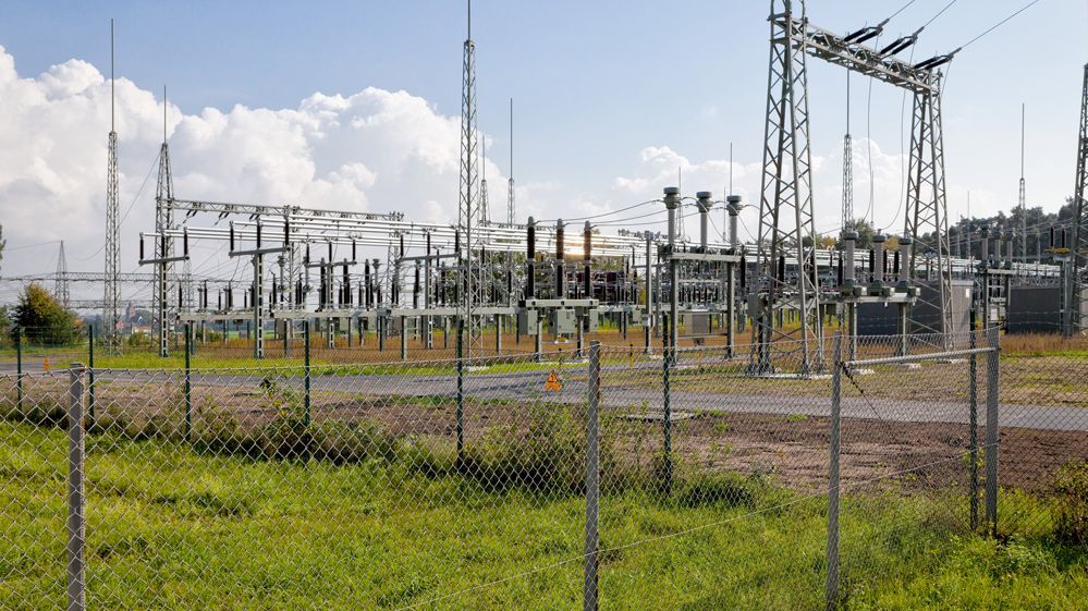 Umspannwerk mit Hochspannungsleitungen, Transformatoren und Metallgerüsten, umgeben von einem Sicherheitszaun an einem sonnigen Tag.