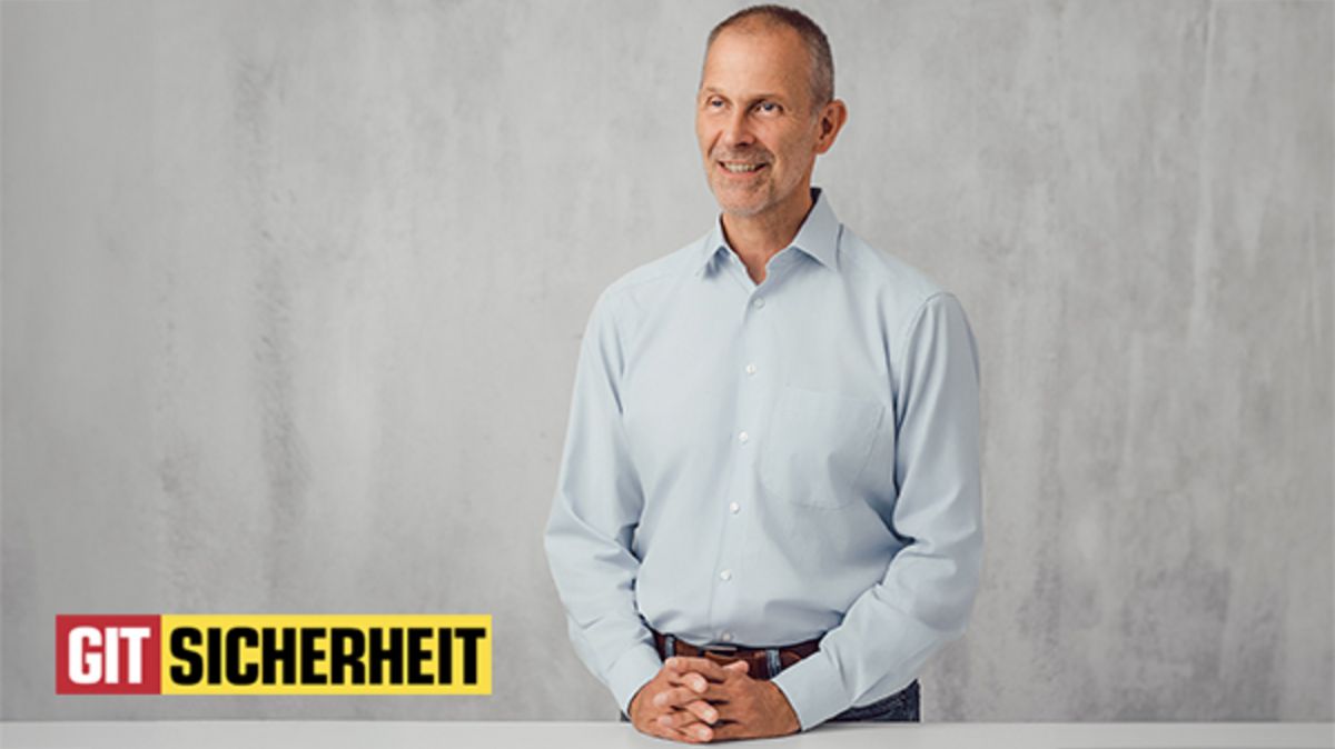 Im Mittelpunkt des Bildes steht Peter Treutler, Prokurist und Leiter der Business Unit IPS bei Securiton, an einem Tisch und vor einer grauen Wand. Links unten ist das "GIT Sicherheit"-Logo zu sehen.