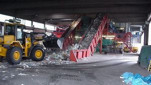 In einer Mülldeponie wird der Müll von einem gelben Bagger auf ein Förderband geladen. 