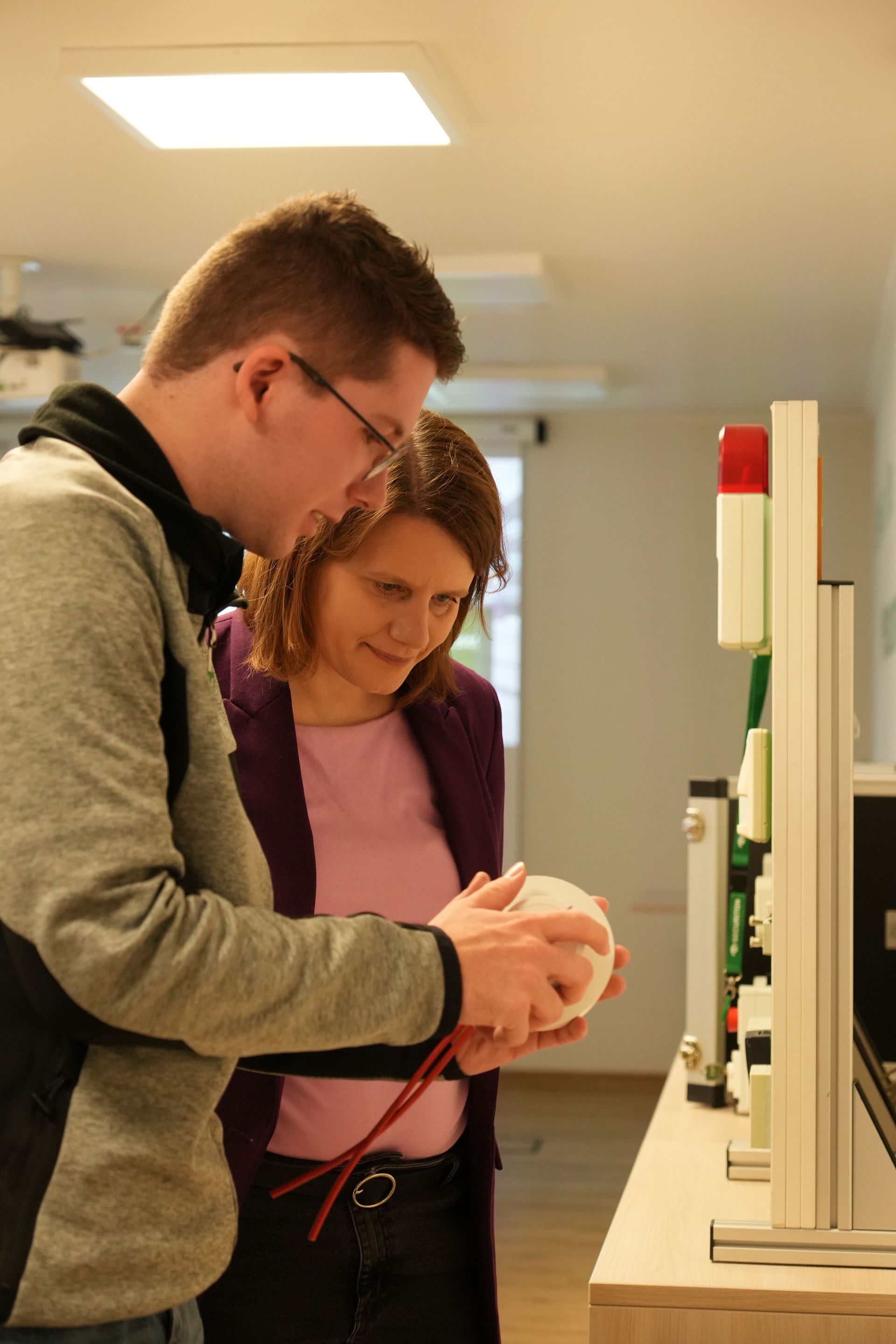Die stellvertretende niedersächsische Ministerpräsidentin Julia Willie Hamburg steht mit einem Mitarbeiter an einem technischen Demonstrationsstand. Der Securiton Mitarbeiter zeigt ihr einen Rauchmelder.