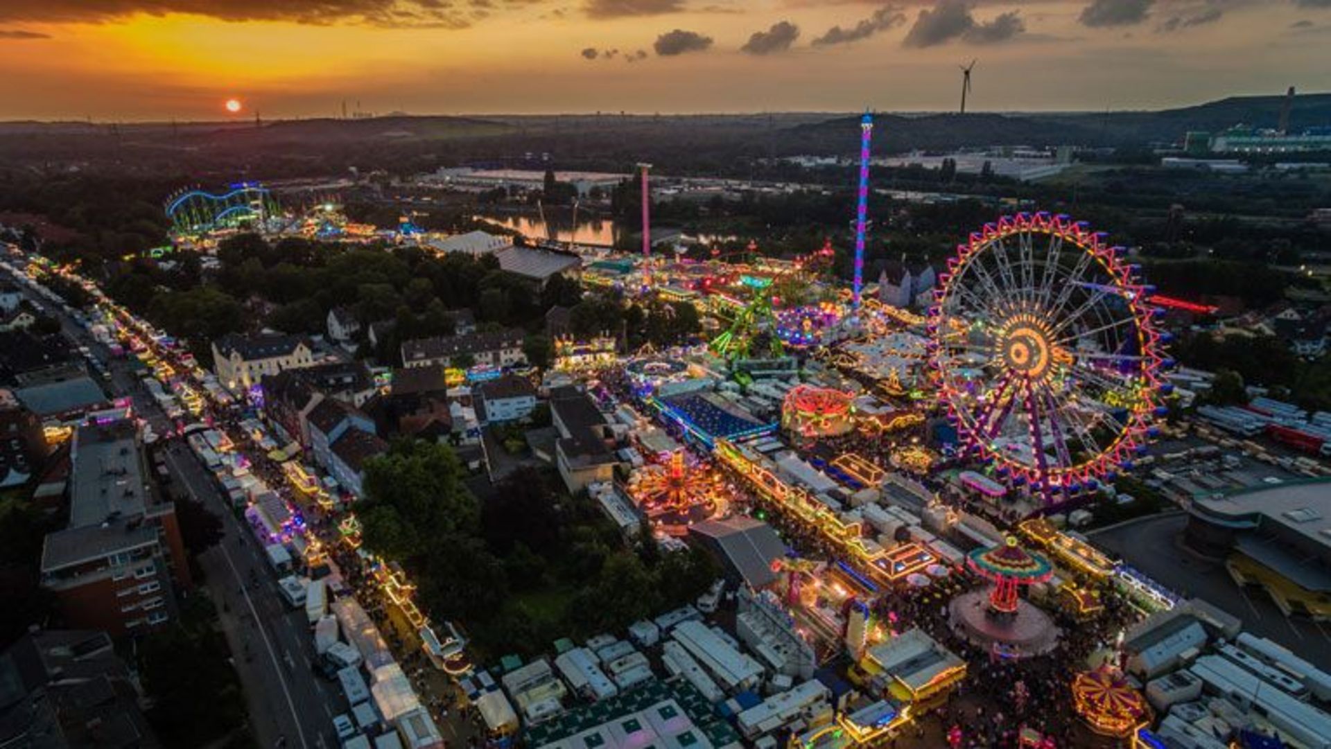 Luftaufnahme eines großen, farbenfrohen Volksfests bei Sonnenuntergang. Zu sehen sind zahlreiche beleuchtete Fahrgeschäfte, darunter ein großes Riesenrad, eine Achterbahn und mehrere Karussells. Dichte Besucherströme bewegen sich durch die Gassen, während Wohnwagen und Festzeltbereiche den äußeren Rand des Geländes säumen. Im Hintergrund erstreckt sich eine weitläufige Stadt- und Industrielandschaft.