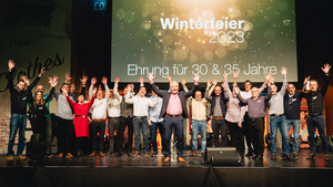 Die Mitarbeiter von Securiton Deutschland, die ihr 2023 ihr Jubiläum feiern stehen gemeinsam auf der Bühne. Im Hintergrund steht auf einer Leinwand "Winterfeier 2023"