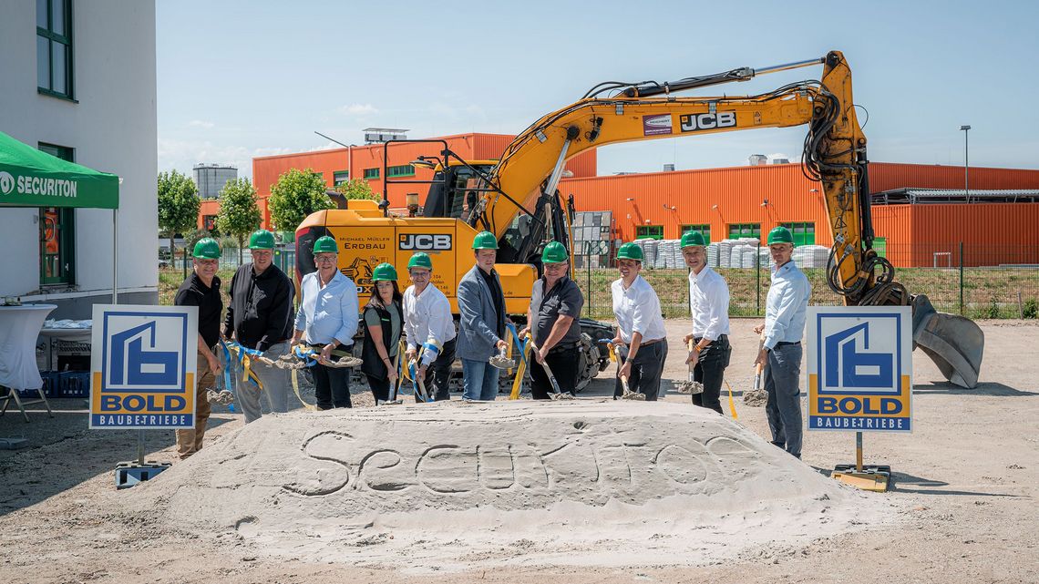Spatenstich für den Securiton-Neubau: Mehrere Personen mit grünen Schutzhelmen stehen mit Spaten vor einem Sandhügel mit der Aufschrift „Securiton“. Im Hintergrund sind ein Bagger und Baustellenanlagen zu sehen.