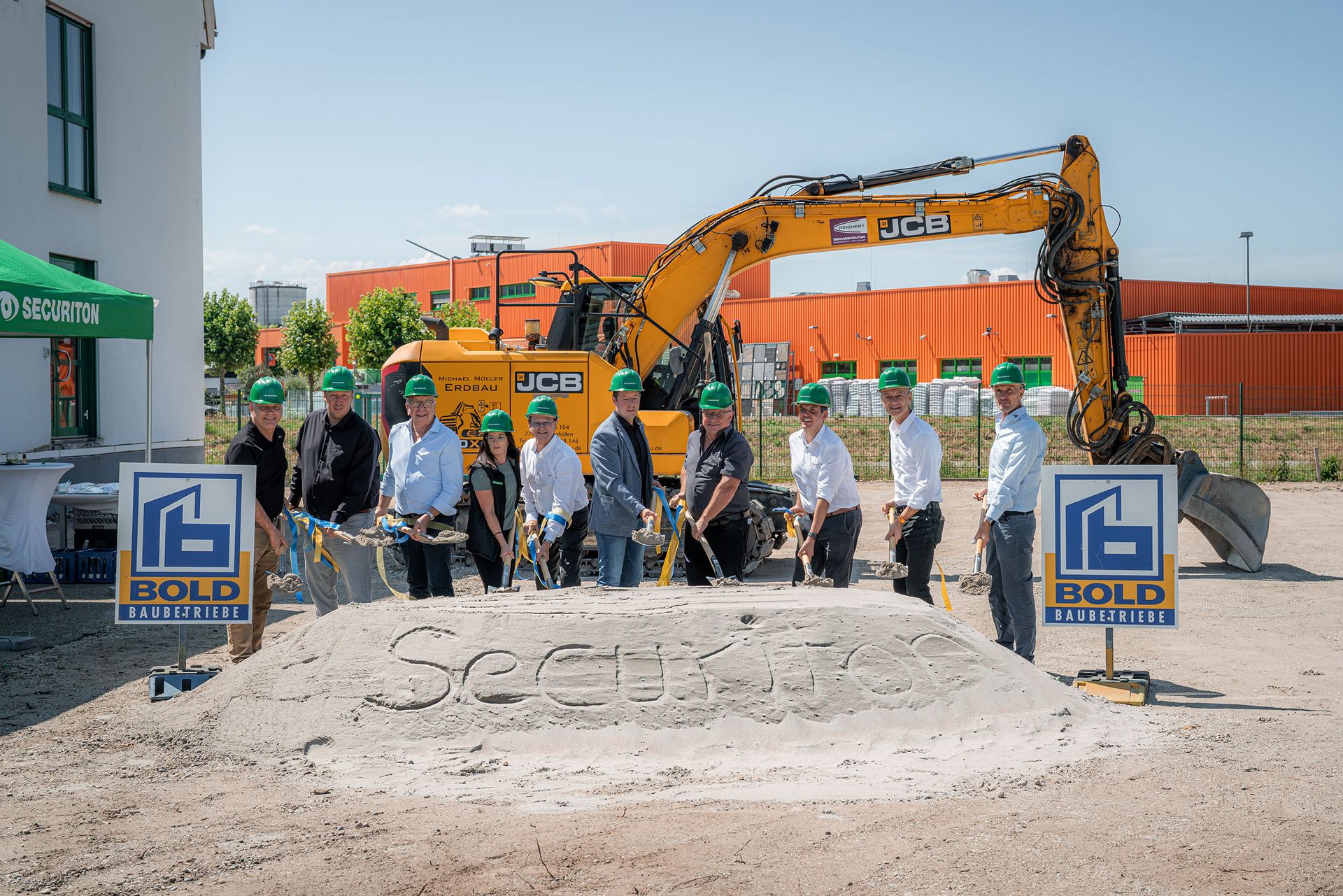 Spatenstich für den Securiton-Neubau: Mehrere Personen mit grünen Schutzhelmen stehen mit Spaten vor einem Sandhügel mit der Aufschrift „Securiton“. Im Hintergrund sind ein Bagger und Baustellenanlagen zu sehen.