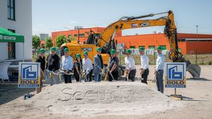 Spatenstich für den Securiton-Neubau: Mehrere Personen mit grünen Schutzhelmen stehen mit Spaten vor einem Sandhügel mit der Aufschrift „Securiton“. Im Hintergrund sind ein Bagger und Baustellenanlagen zu sehen.