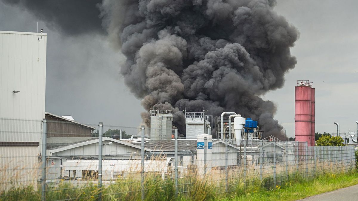 Dichter schwarzer Rauch steigt aus einer Industrieanlage auf. Das Brandereignis findet auf einem umzäunten Betriebsgelände mit mehreren Produktions- und Lagereinrichtungen statt. Im rechten Bildbereich sind Einsatzfahrzeuge der Feuerwehr zu sehen. Die starke Rauchentwicklung verdeutlicht die Auswirkungen eines Großbrands in einem industriellen Umfeld.
