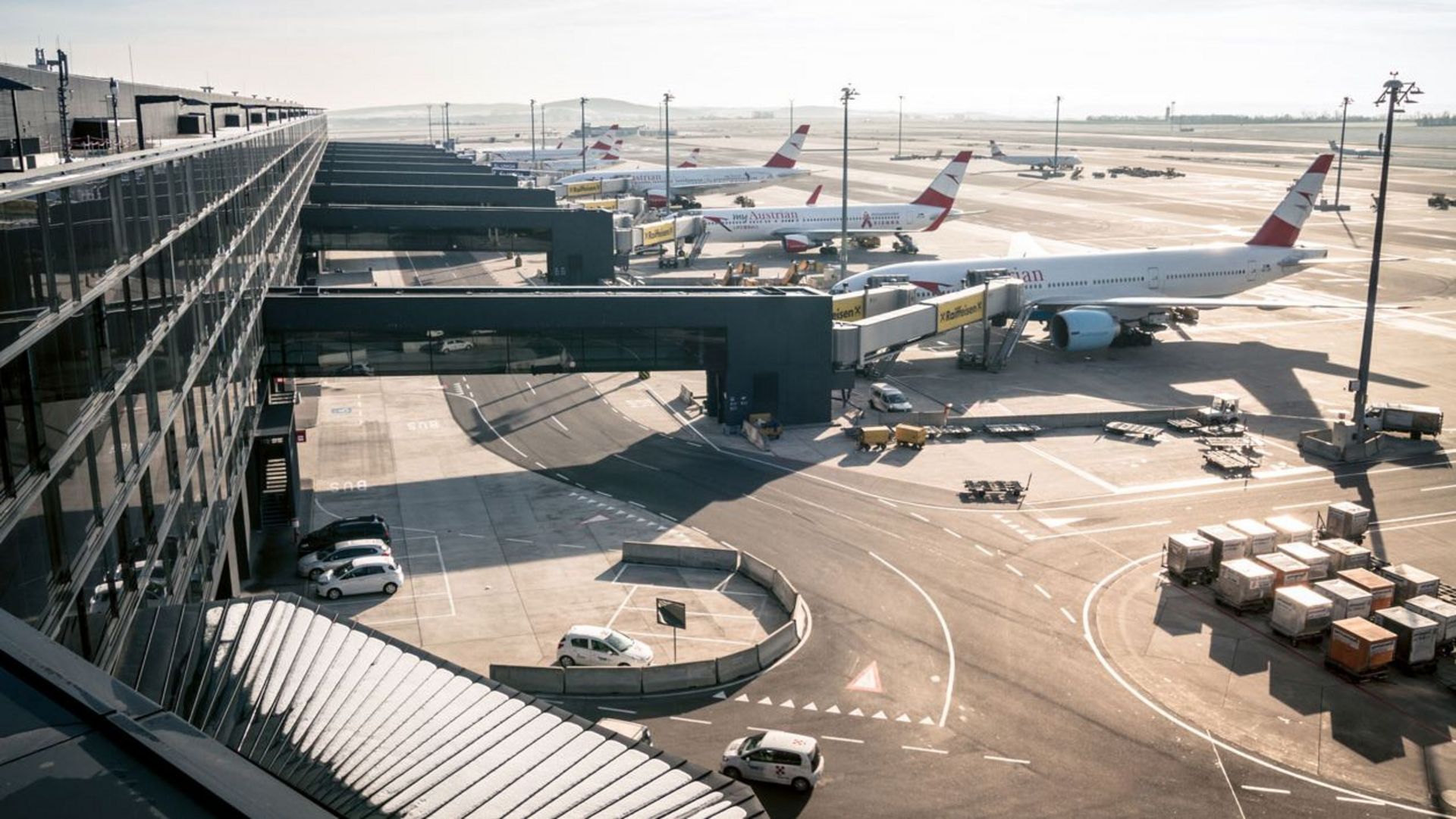 Blick von einem Terminalgebäude auf ein Flughafenvorfeld mit mehreren abgestellten und angedockten Passagierflugzeugen. Mehrere Fluggastbrücken führen vom Terminal zu den Flugzeugen. Auf dem Vorfeld befinden sich Servicefahrzeuge, Container und Abfertigungsequipment. Die Szene ist bei Tageslicht mit klarer Sicht aufgenommen.“