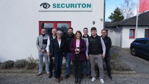 Gruppenfoto vor dem Firmengebäude von Securiton Deutschland in Wunstorf. In der Mitte steht die stellvertretende niedersächsische Ministerpräsidentin Julia Willie Hamburg, die ein kleines Stoffschaf in den Händen hält. Um sie herum stehen sieben weitere Personen aus dem Unternehmensumfeld.