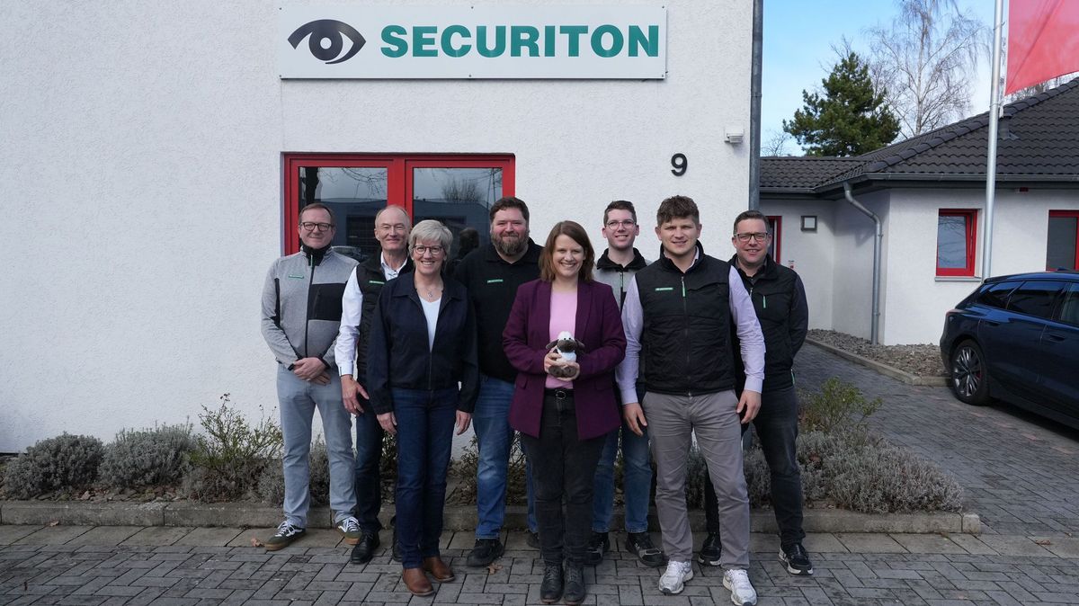Gruppenfoto vor dem Firmengebäude von Securiton Deutschland in Wunstorf. In der Mitte steht die stellvertretende niedersächsische Ministerpräsidentin Julia Willie Hamburg, die ein kleines Stoffschaf in den Händen hält. Um sie herum stehen sieben weitere Personen aus dem Unternehmensumfeld.