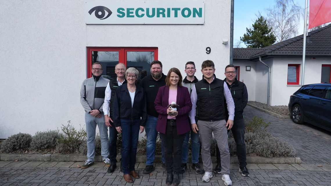 Gruppenfoto vor dem Firmengebäude von Securiton Deutschland in Wunstorf. In der Mitte steht die stellvertretende niedersächsische Ministerpräsidentin Julia Willie Hamburg, die ein kleines Stoffschaf in den Händen hält. Um sie herum stehen sieben weitere Personen aus dem Unternehmensumfeld.