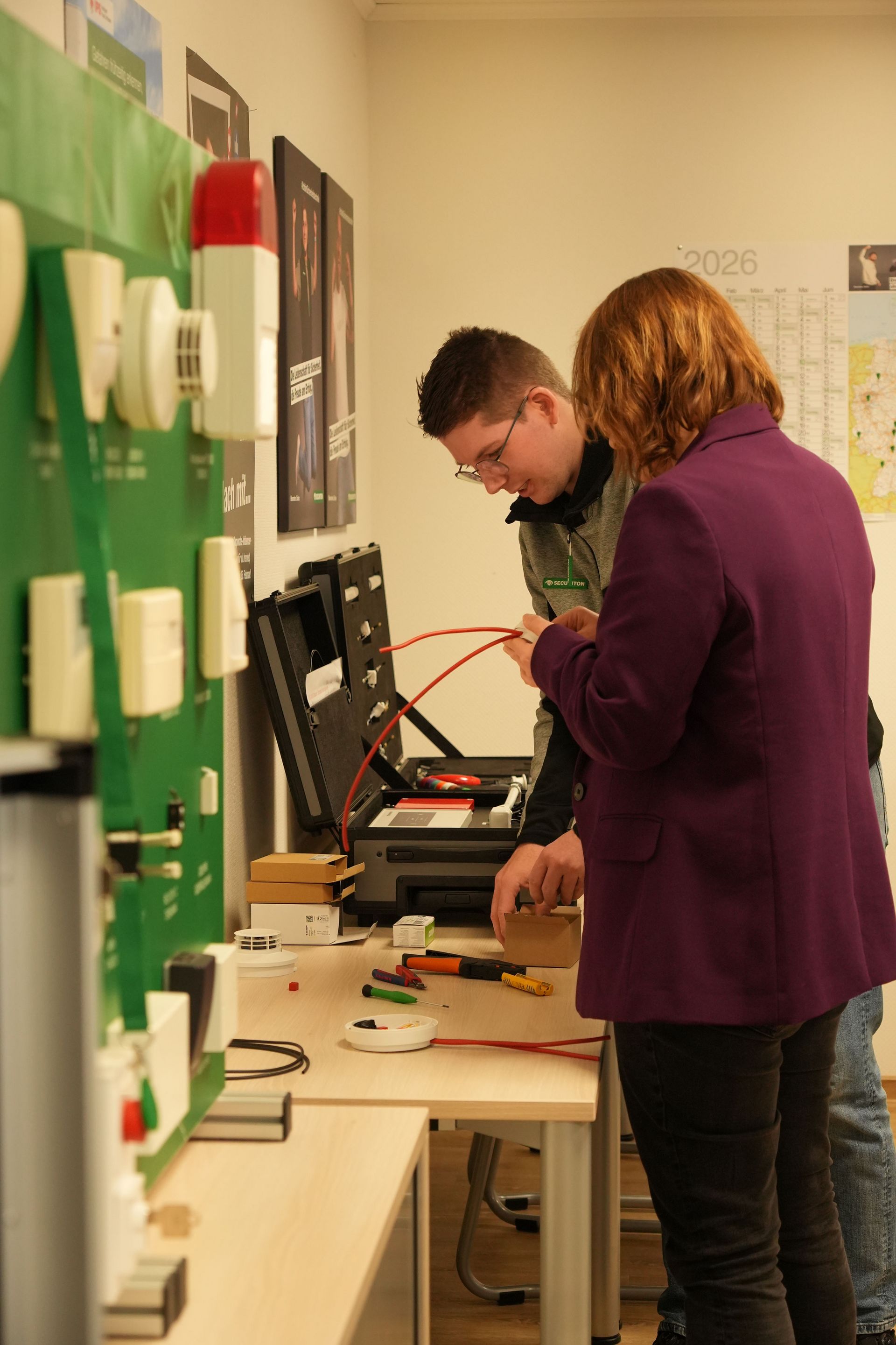 Die stellvertretende niedersächsische Ministerpräsidentin Julia Willie Hamburg steht mit einem Mitarbeiter von Securiton Deutschland an einem Arbeitstisch und betrachtet technische Geräte und Kabel. Der Mitarbeiter zeigt ihr ein rotes Kabel aus einem offenen Werkzeugkoffer. Im Vordergrund sind weitere technische Komponenten zu sehen.