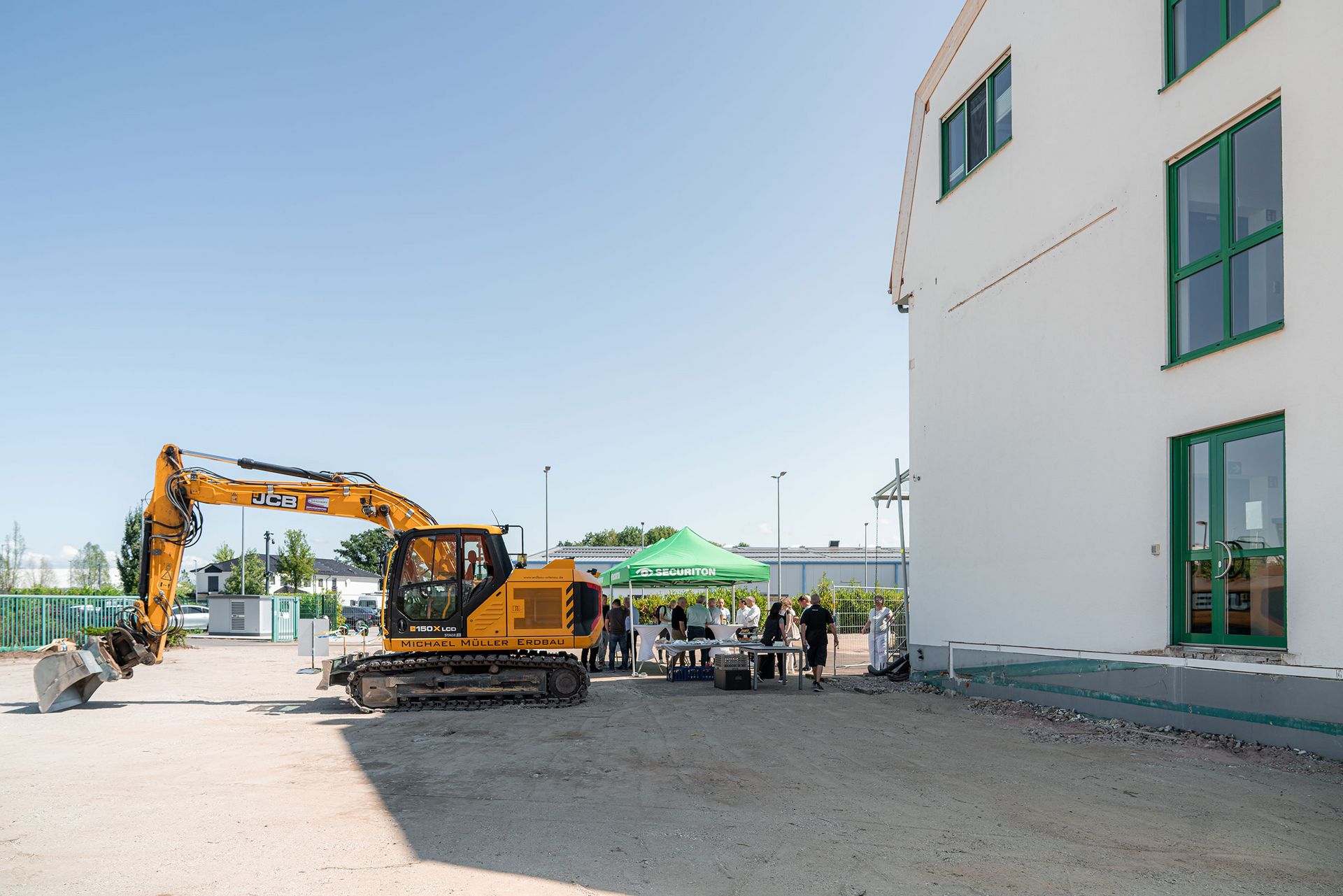 Baustellenszene auf dem Securiton-Gelände mit gelbem Bagger, einem Securiton-Pavillon und Gästen vor einem Bestandsgebäude während einer Neubauveranstaltung.
