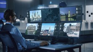 Ein Mann sitzt an einem Schreibtisch und schaut auf sechs Monitore die Aufnahmen von verschiedenen Städten und deren Sehenswürdigkeiten zeigen (z.B. London Big Ben, Italien Koloseum)